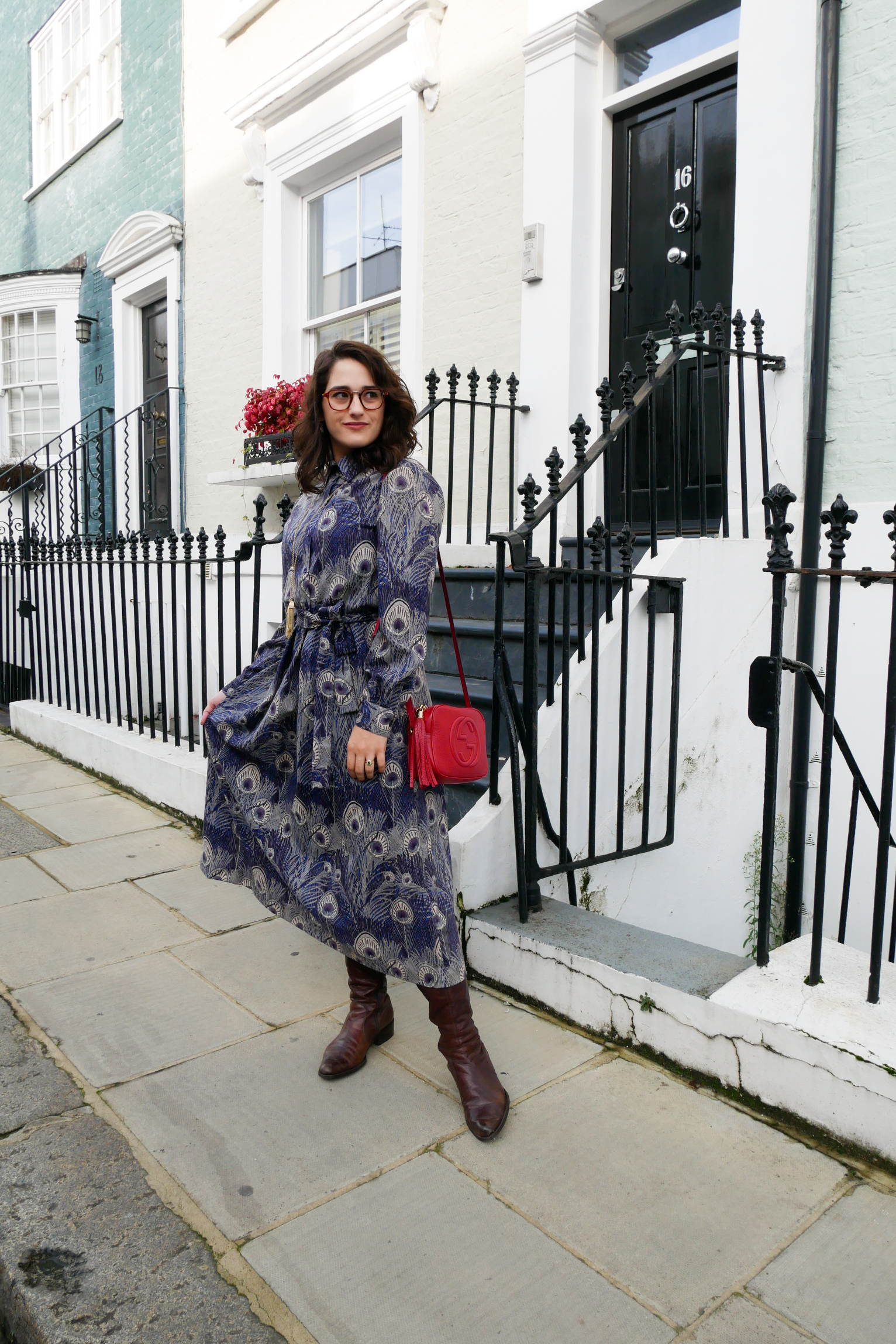 Katie wears a purple and blue peacock print midi length shirt dress with calf high brown boots, a red shoulder bag and gold tone tassel necklace on one of Notting Hill's rainbow house-front streets.