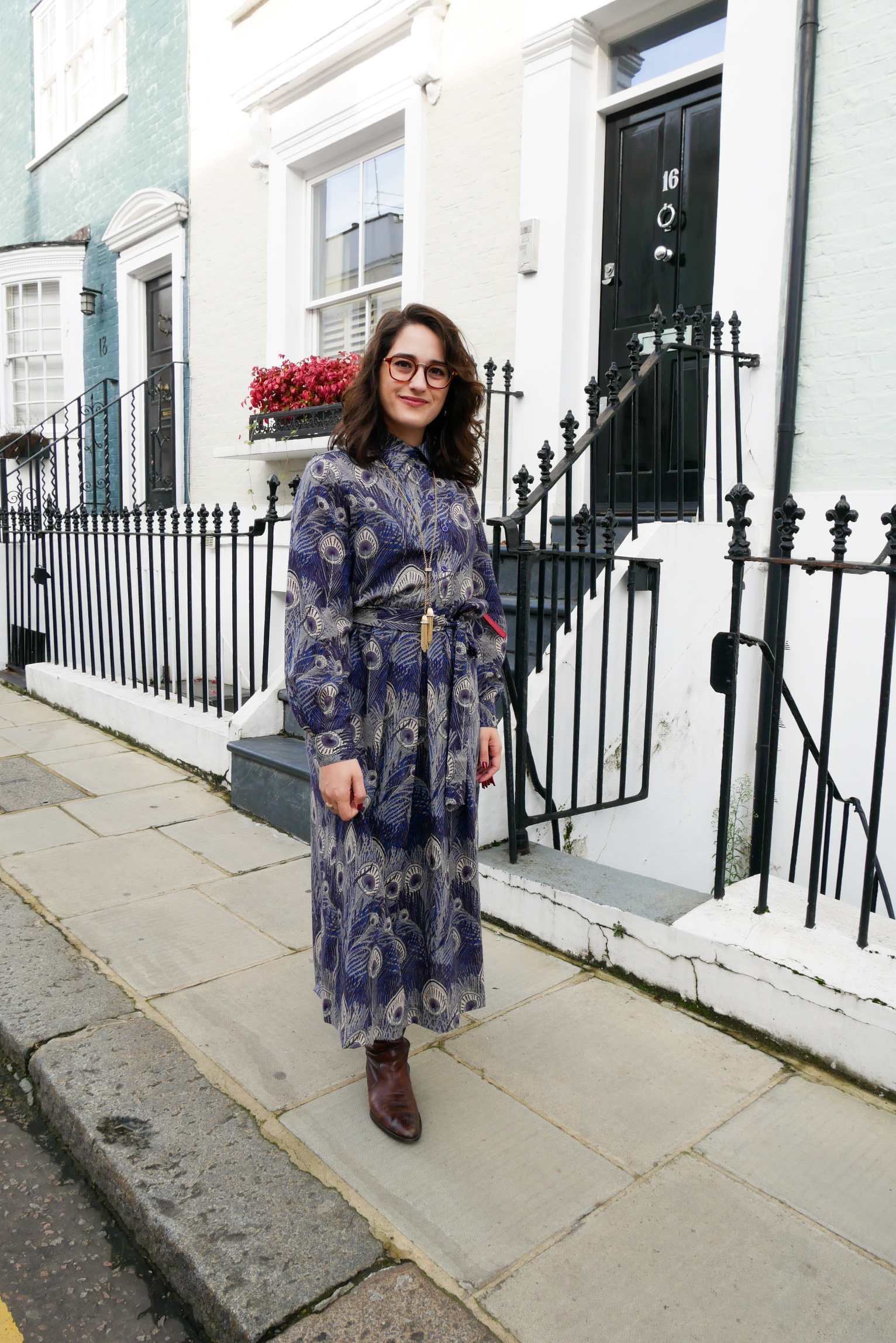 Katie wears a purple and blue peacock print midi length shirt dress with calf high brown boots, a red shoulder bag and gold tone tassel necklace on one of Notting Hill's rainbow house-front streets.