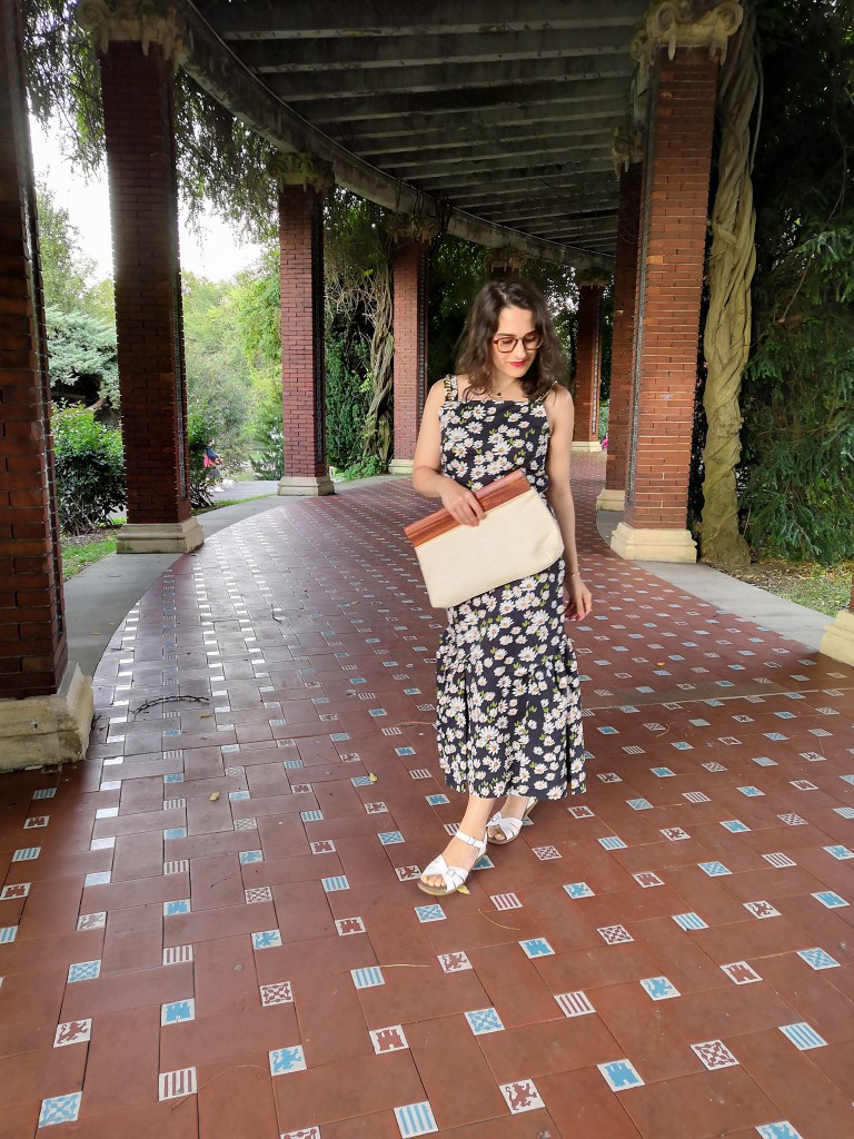 Katie wears a maxi dress with a daisy print and chains on the straps, white sandals and a cream cotton and wooden handled clutch bag. She's posing in a Spanish park.