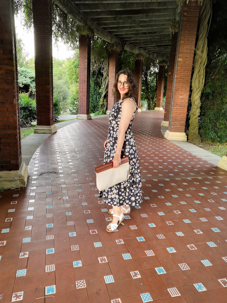 Katie wears a maxi dress with a daisy print and chains on the straps, white sandals and a cream cotton and wooden handled clutch bag. She's posing in a Spanish park.