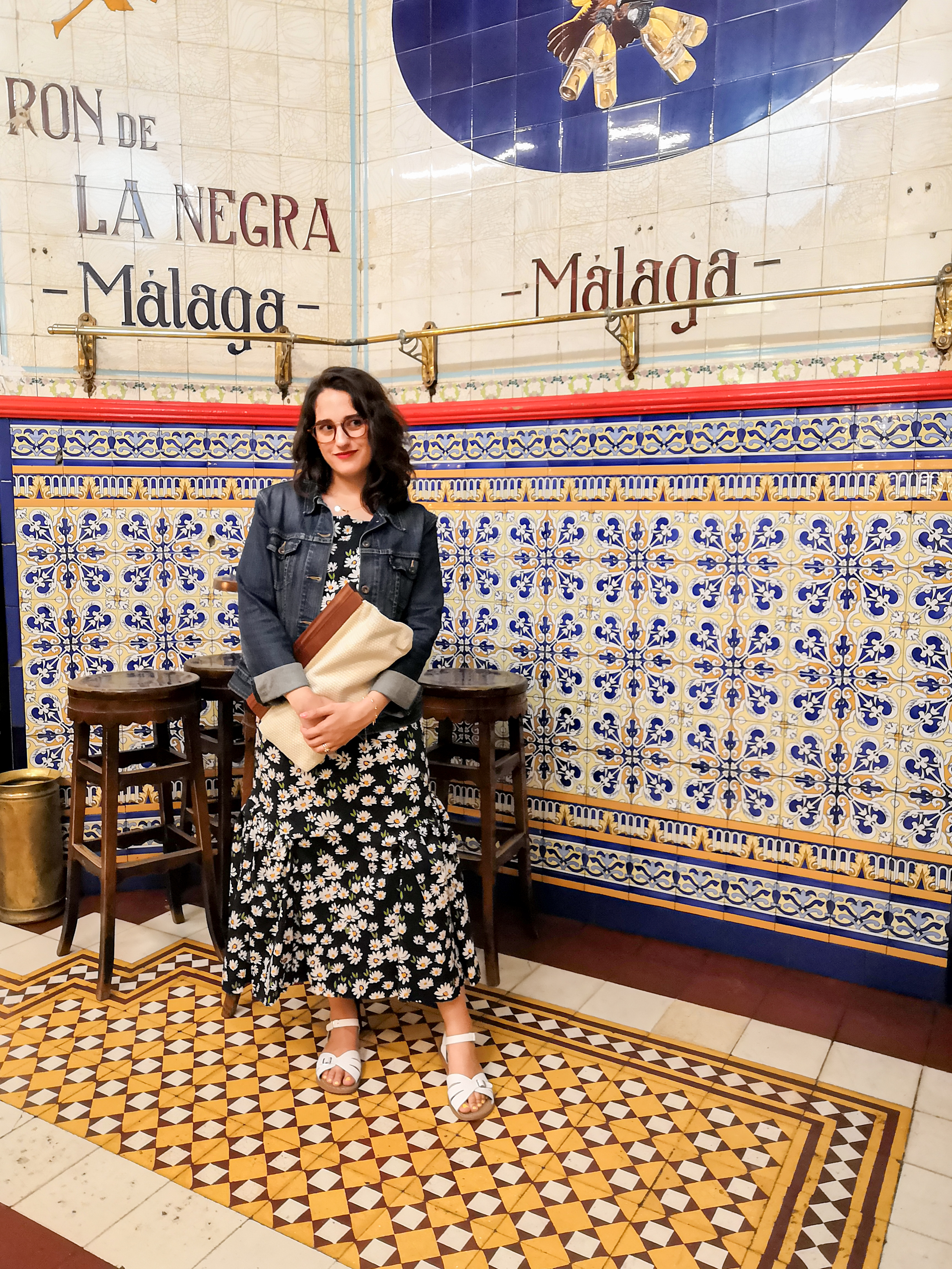 Katie wears a maxi dress with a daisy print and chains on the straps, white sandals,  a cream cotton and wooden handled clutch bag and a denim jacket with cuffed sleeves. She's standing in a Spanish bar with tiled floors and walls.