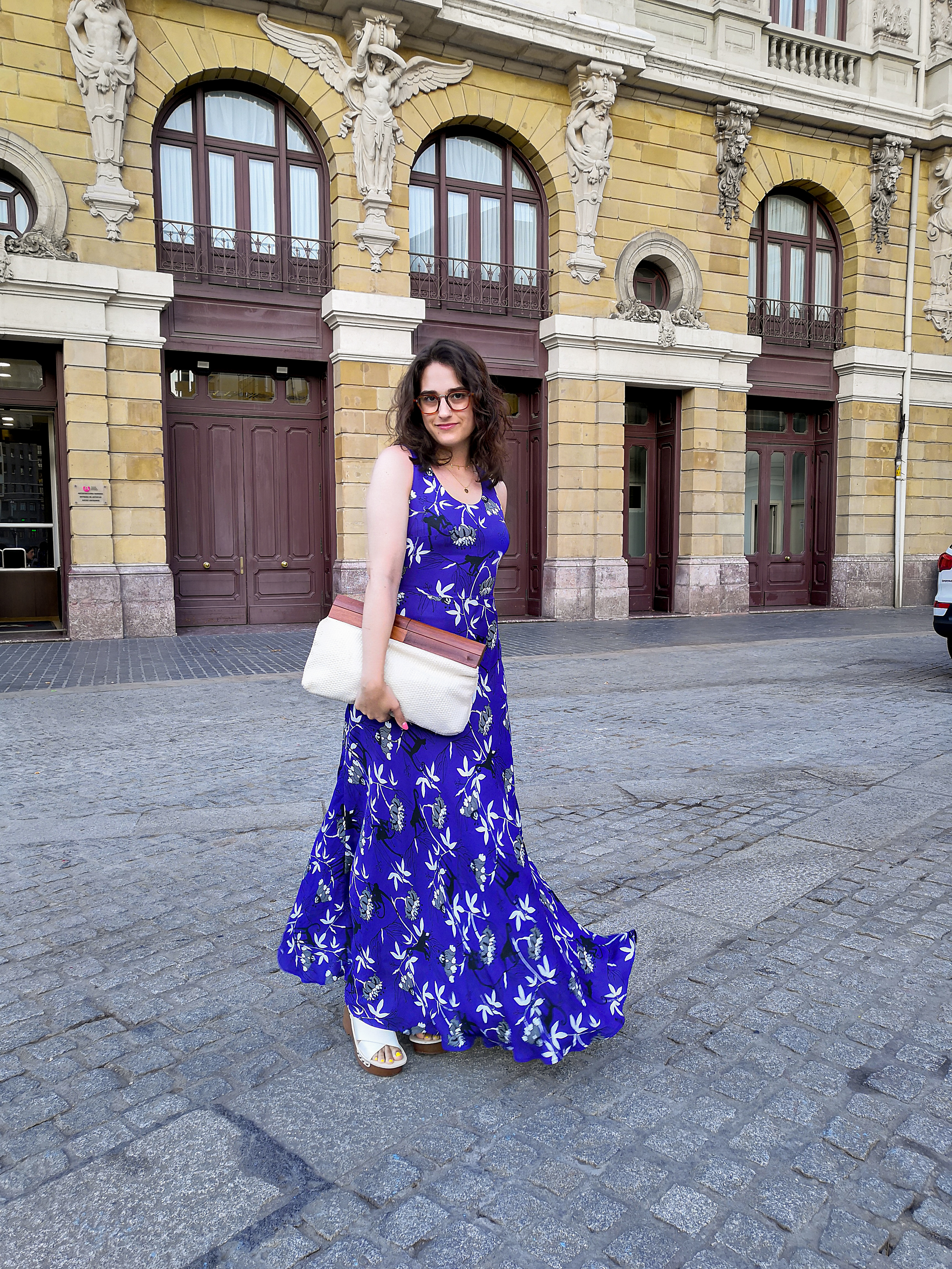 Katie wears a dark blue sleeveless maxi dress with a monkey and palm tree print. She's holding a white cotton clutch bag with a wooden handle, and wooden platform sandals with a cream leather upper. She's standing on a side street in Bilbao with a large old building behind her.