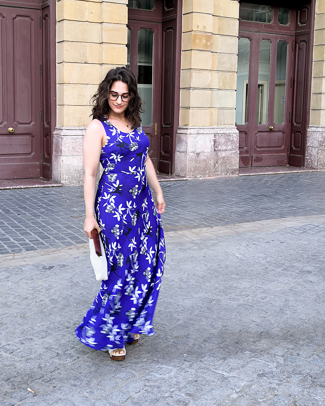 Katie wears a dark blue sleeveless maxi dress with a monkey and palm tree print. She's holding a white cotton clutch bag with a wooden handle, and wooden platform sandals with a cream leather upper. She's standing on a side street in Bilbao with a large old building behind her.