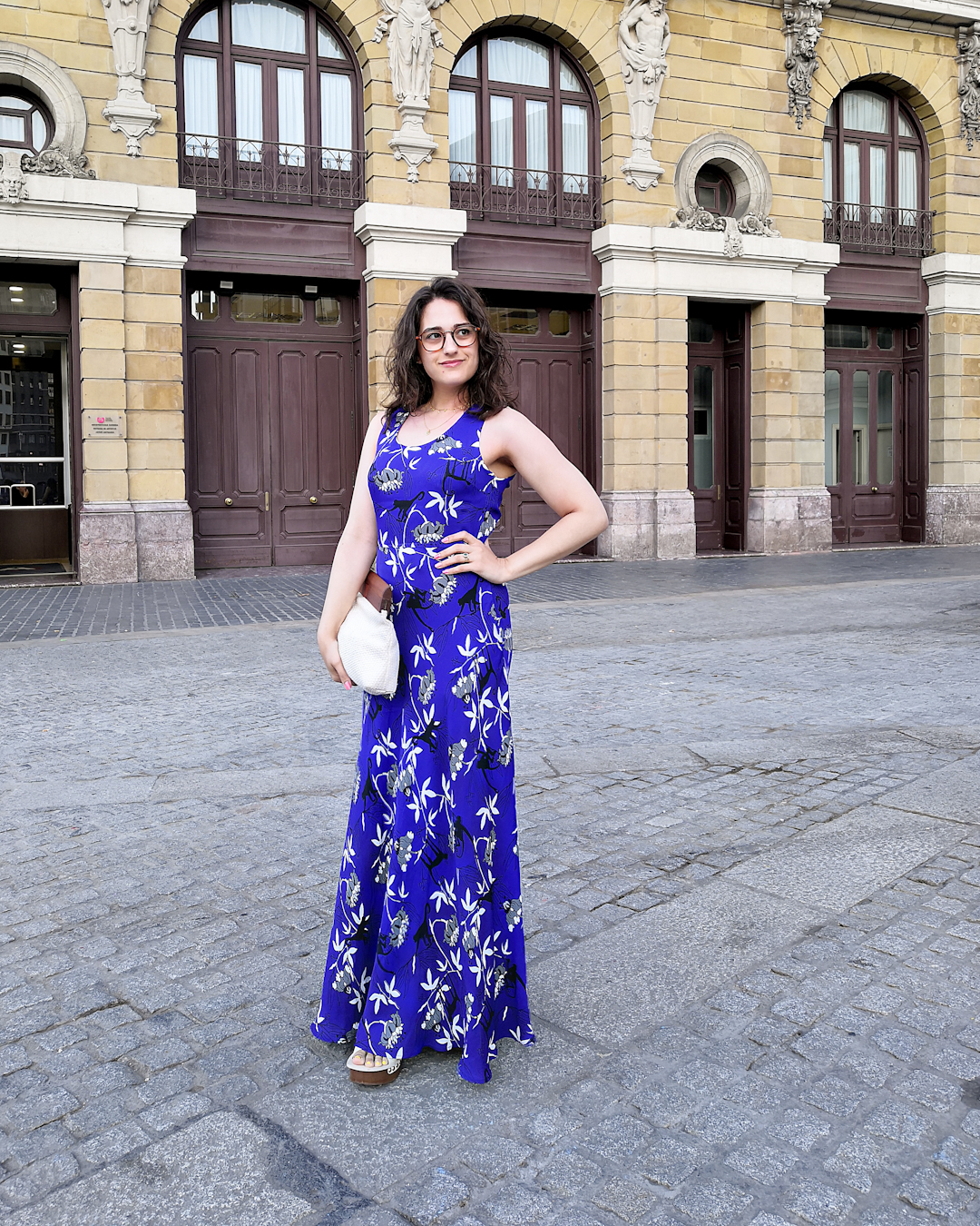 Katie wears a dark blue sleeveless maxi dress with a monkey and palm tree print. She's holding a white cotton clutch bag with a wooden handle, and wooden platform sandals with a cream leather upper. She's standing on a side street in Bilbao with a large old building behind her.