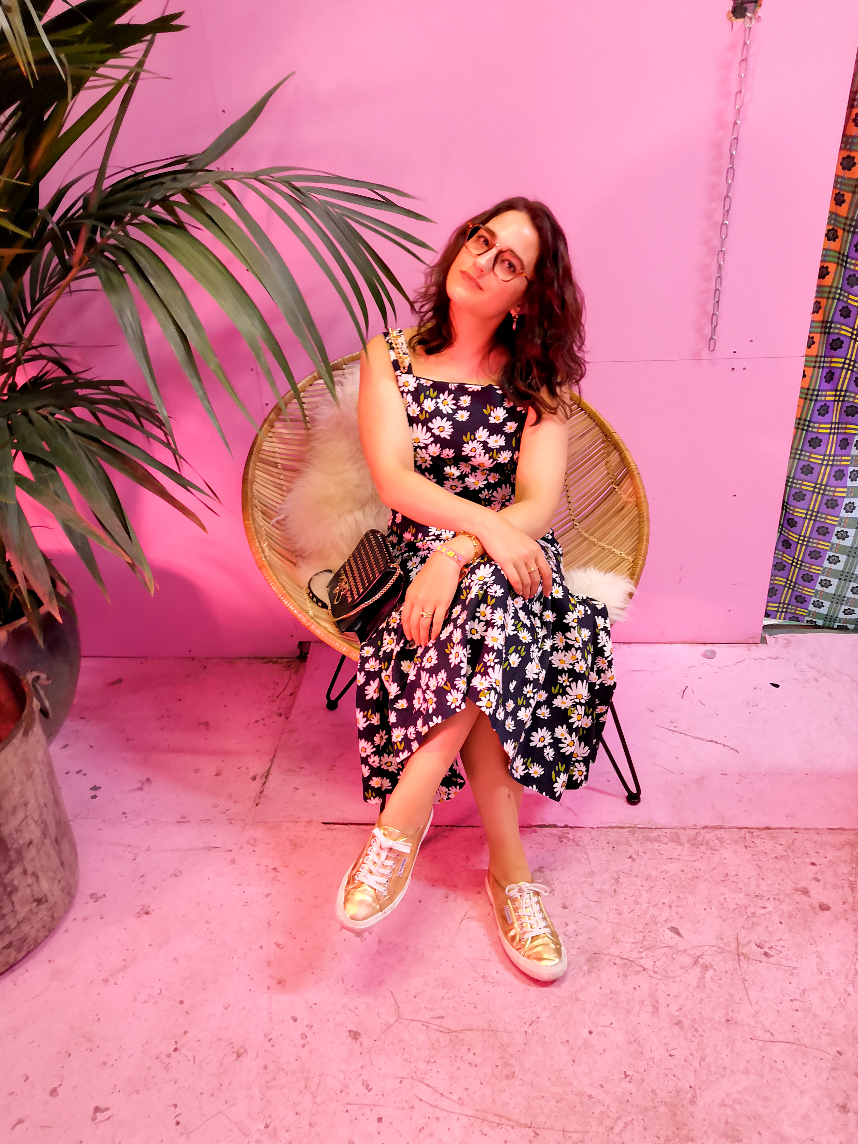Katie wears a black and white floral maxi dress. She sits on a wicker chair next to a large potted plant in a room with pink walls.