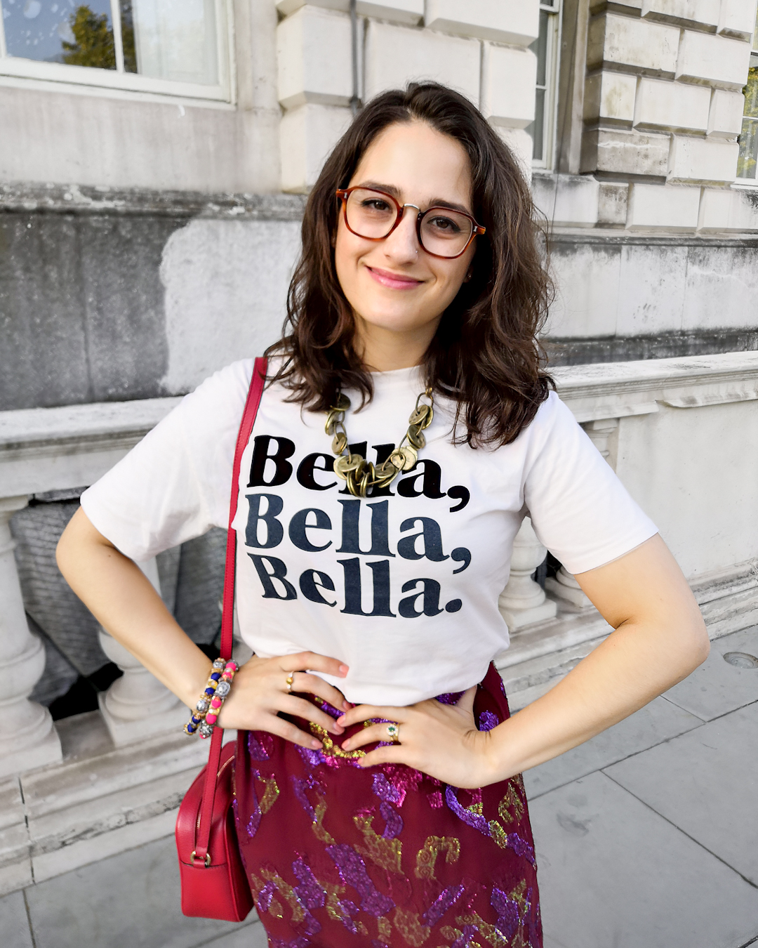Katie wears a cream T shirt with the words "Bella Bella Bella" printed in navy serif font, an assymeyric cranberry maxi skirt with a green, purple and fuschia metallic print, and slip on pink shoes. She's accessorised with a brass layered necklace and multi colour Pakistani bangles.