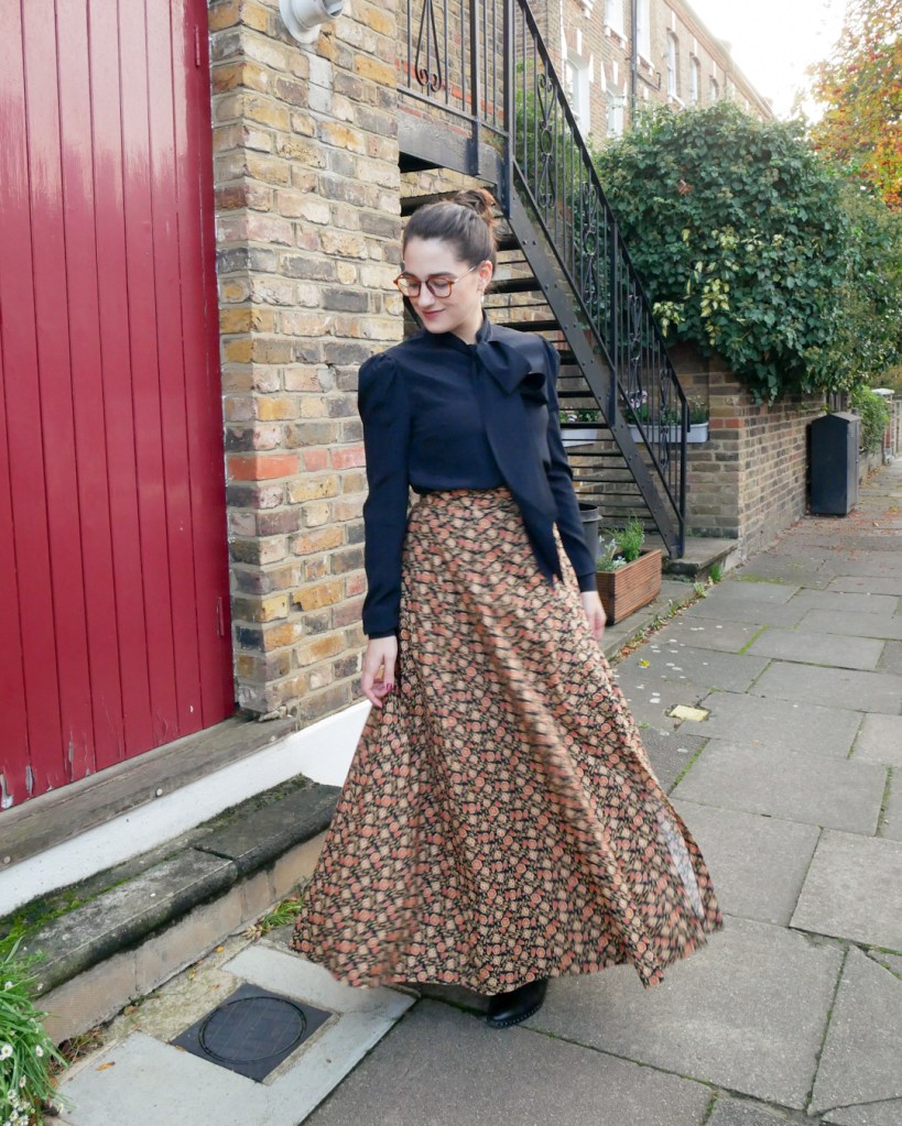 Katie wears a silky black blouse with long sleeves and a pussybow at the neck, matched with an autumnal coloured maxi skirt with ditsy floral print and black ankle boots.