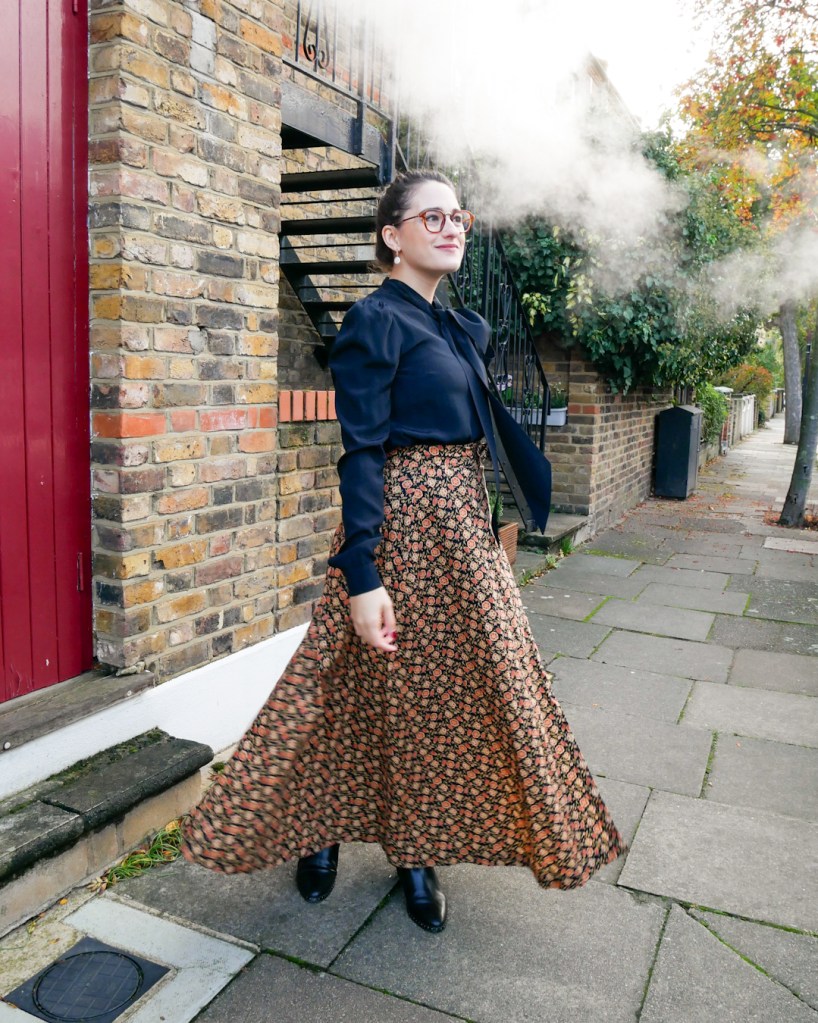 Katie wears a silky black blouse with long sleeves and a pussybow at the neck, matched with an autumnal coloured maxi skirt with ditsy floral print and black ankle boots.
