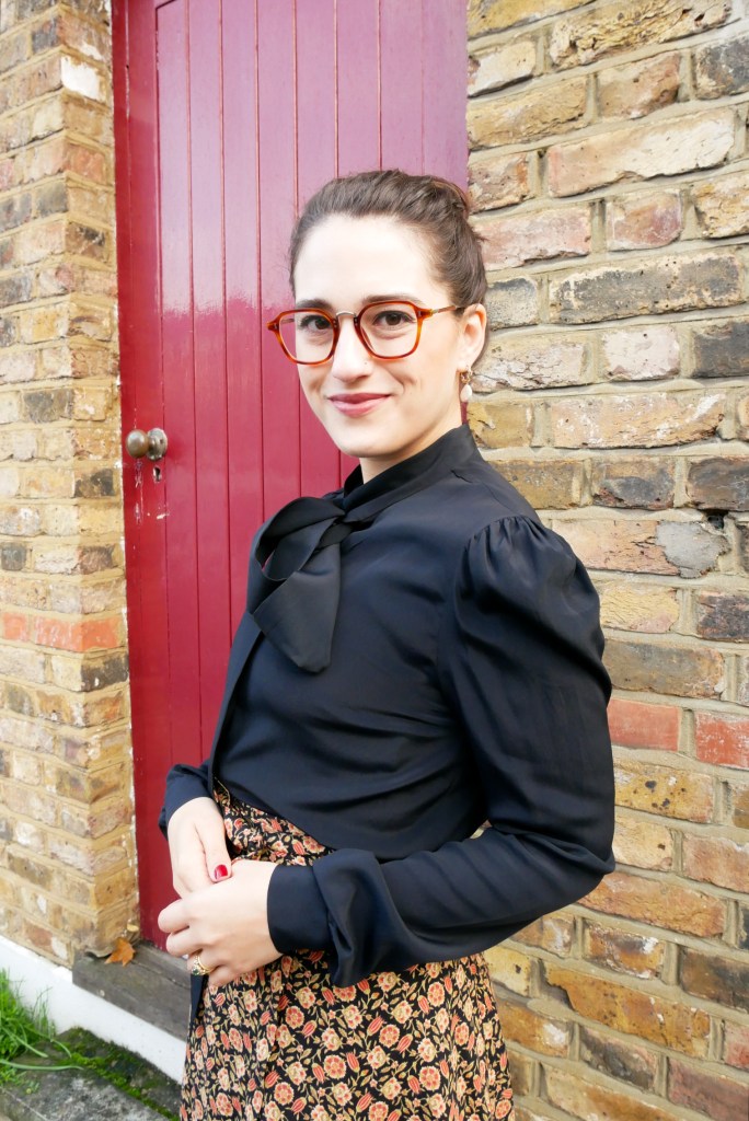 Katie wears a silky black blouse with long sleeves and a pussybow at the neck, matched with an autumnal coloured maxi skirt with ditsy floral print and black ankle boots.
