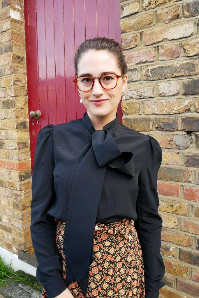 Katie wears a silky black blouse with long sleeves and a pussybow at the neck, matched with an autumnal coloured maxi skirt with ditsy floral print and black ankle boots.