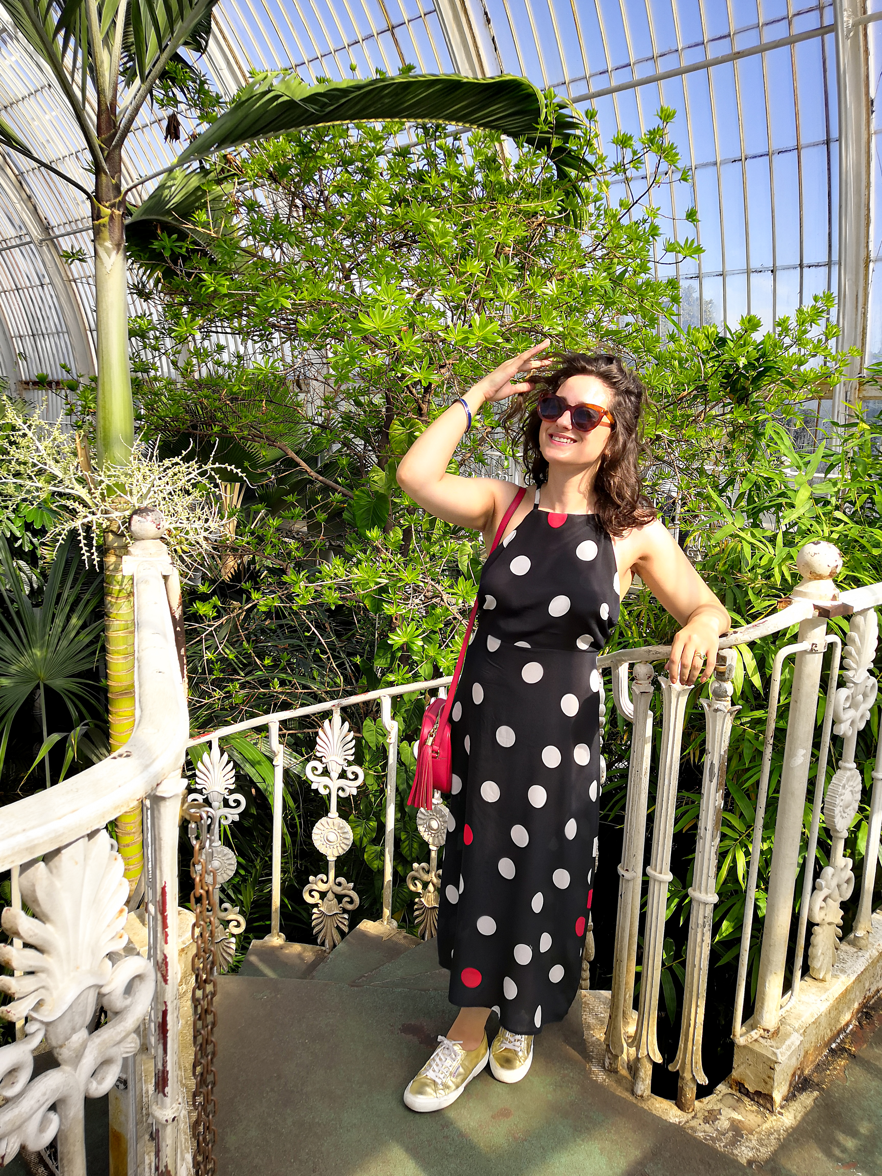 Katie is wearing a navy, white and red statement polkadot maxi dress with spaghetti straps and an open back. The photos have been taken in Kew Gardens on a sunny day and she's surrounded by plants.