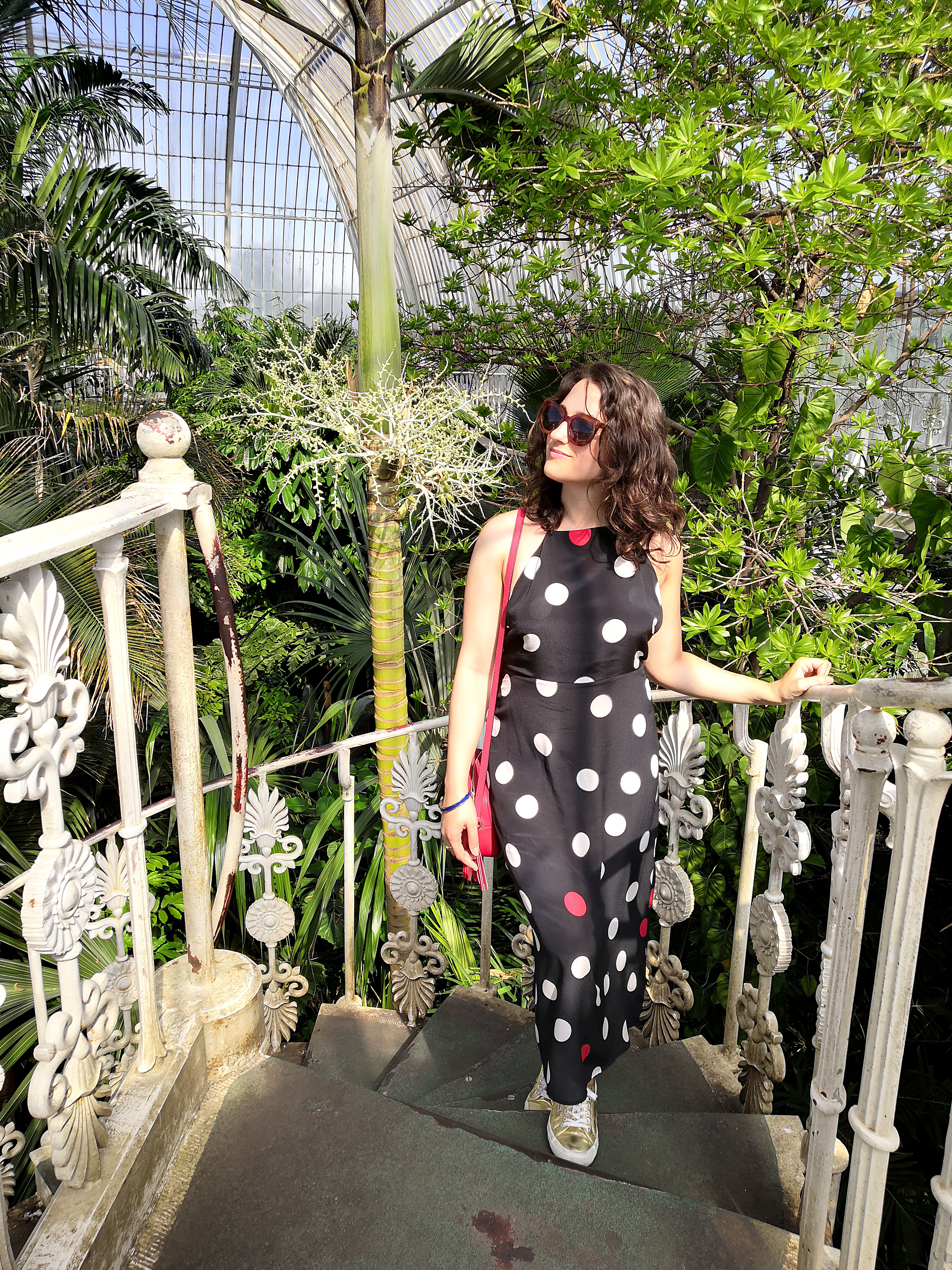 Katie is wearing a navy, white and red statement polkadot maxi dress with spaghetti straps and an open back. The photos have been taken in Kew Gardens on a sunny day and she's surrounded by plants.