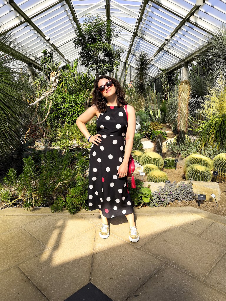 Katie is wearing a navy, white and red statement polkadot maxi dress with spaghetti straps and an open back. The photos have been taken in Kew Gardens on a sunny day and she's surrounded by plants.