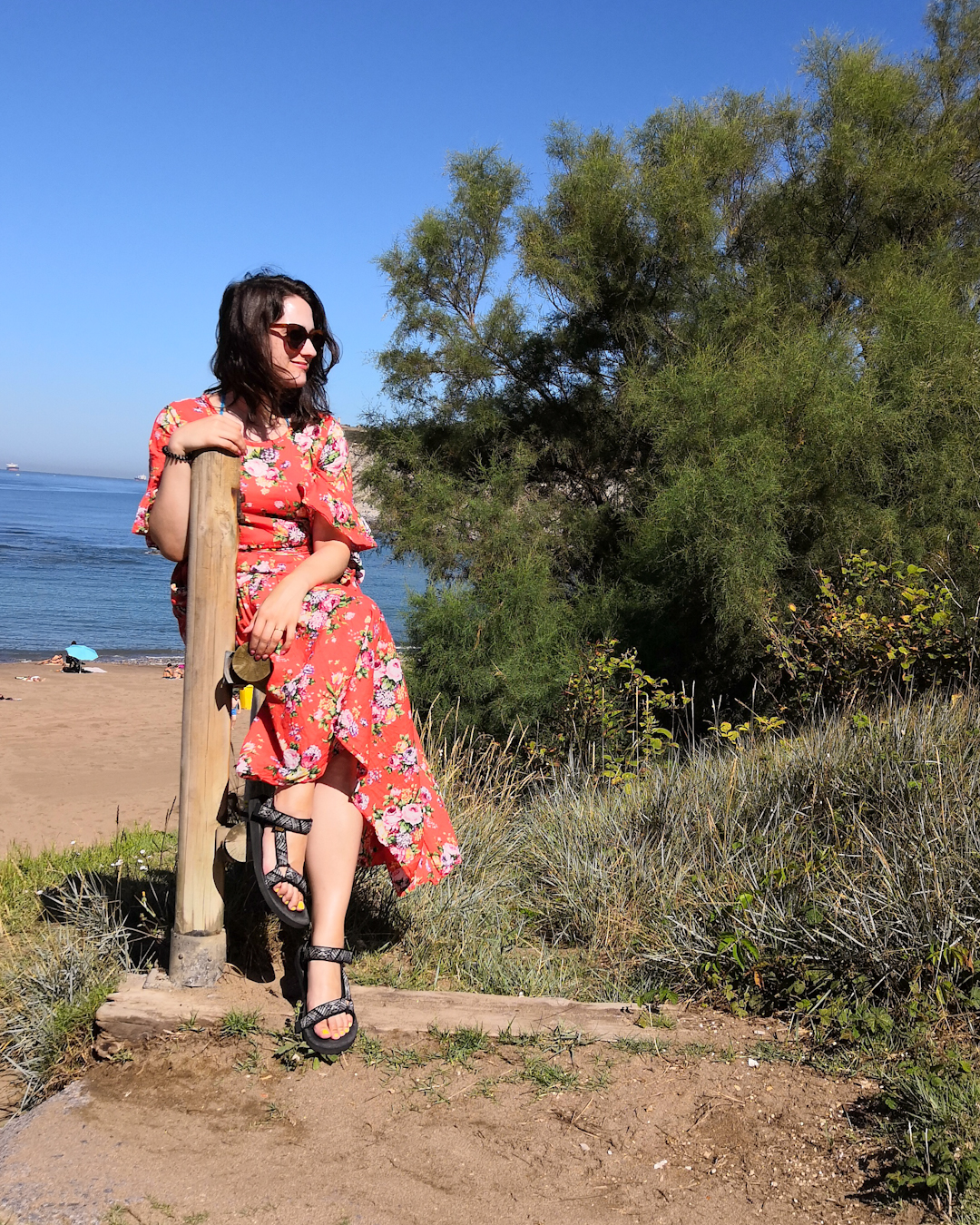 Katie is at the beach. She wears sunglasses & an orange maxi dress with floral pattern & tiered hem. She sits on a wooden rail leading to the beach, the sea is behind her.