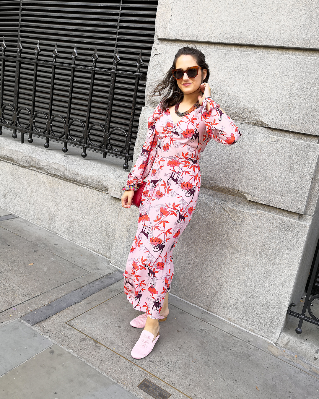 Katie wears a pink maxi dress with fuschia flowers and brown monkeys printed on. She's also wearing oversized amber coloured cat eye sunglasses, a layered multicoloured pink bugle bead necklace and bangles in the traditional Pakistan craft style.