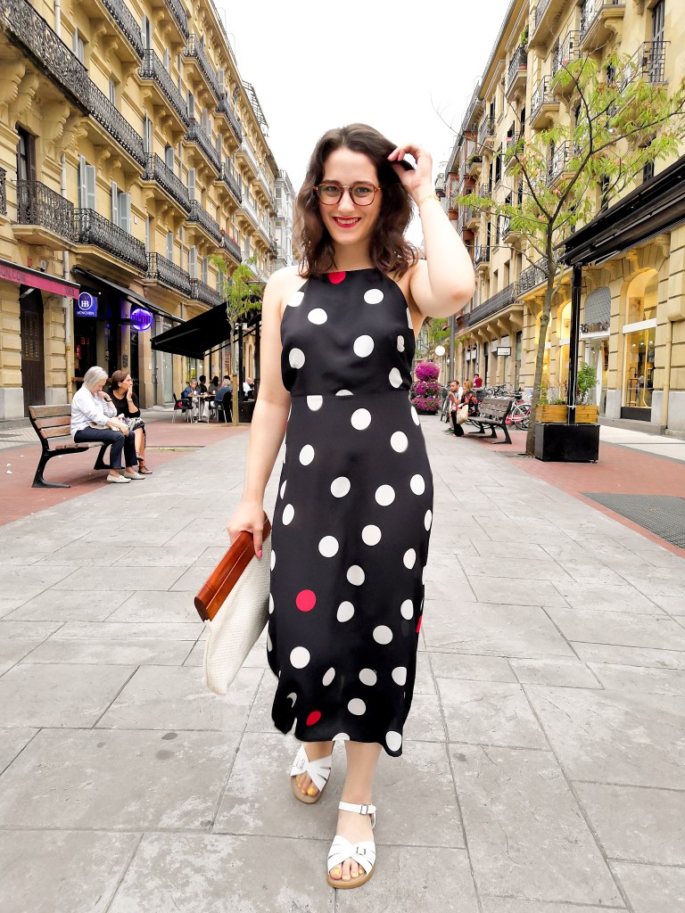 Katie walks towards the camera down a pedestrianised street in Donastia. She's wearing a halter top maxi dress with large polka dots, white sandals, a wood and whiten woven clutch bag and has bright red lipstick.