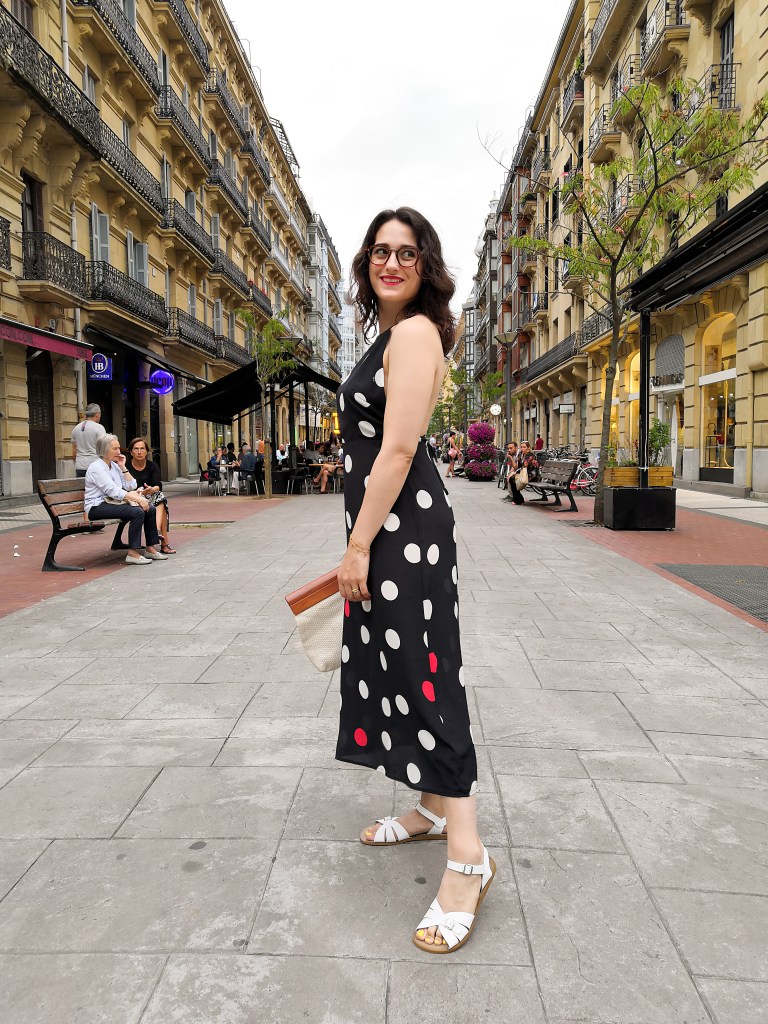 Katie walks towards the camera down a pedestrianised street in Donastia. She's wearing a halter top maxi dress with large polka dots, white sandals, a wood and whiten woven clutch bag and has bright red lipstick.