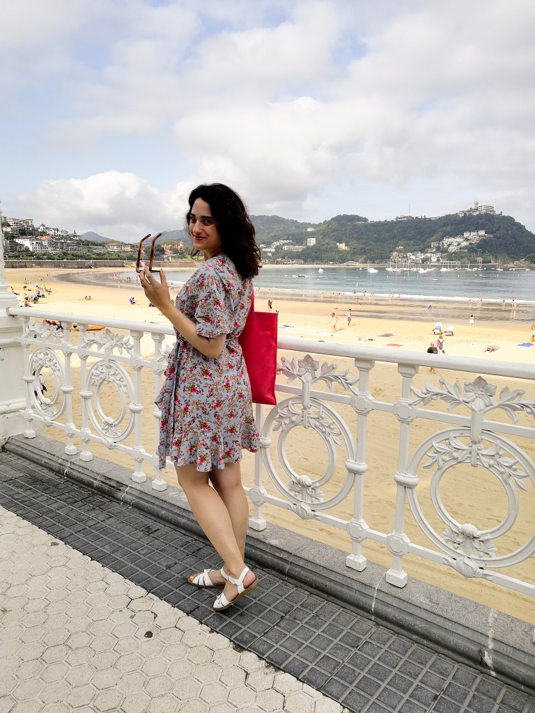 In Donasti on Monte Urgull, Katie sits on a viewing point looking out to the see with her face in profile. Her hair is back and she wears a blue floral wrap dress and white sandals. In the 2nd and 3rd pictures she wears the same outfit at the beach with her hair down.