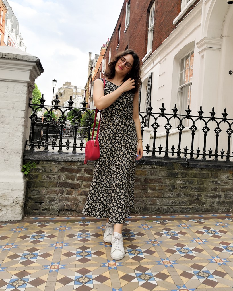 Katie wears a silk maxi dress with a black and white small floral print. It has a thin matching belt and she's paired it with a red handbag and white sneakers. 