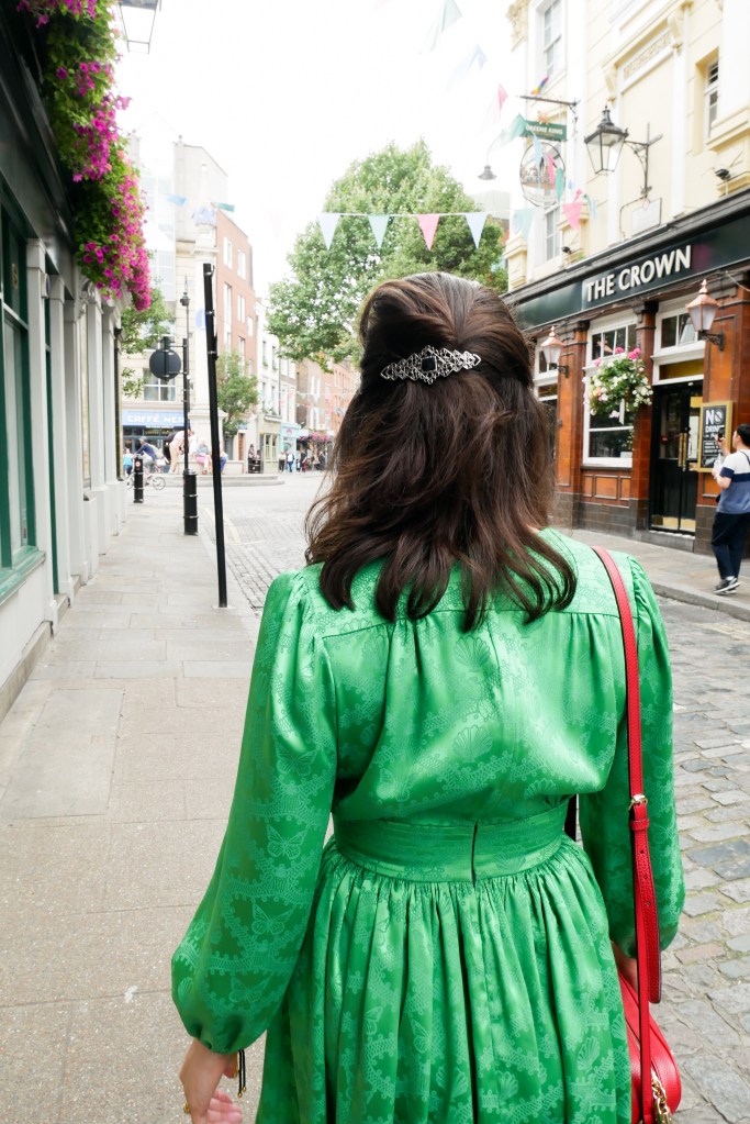 Katie is in Covent Garden, walking away from the camera She wears a green dress with long poofy sleeves, a nipped in waist and short flouncy skirt. She's carrying a red shoulder bag, and a silver and black barrette in her hair.