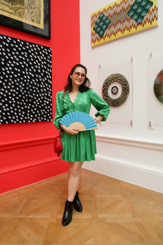 Katie is in the galleries of the Royal Academy Summer Show, where the walls and spaces are crowded with paintings, sculptures, and other artworks. She wears a green dress with long poofy sleeves, a nipped in waist and short flouncy skirt. She's carrying a red shoulder bag and a wood and blue fabric folding fan and wearing black ankle boots.
