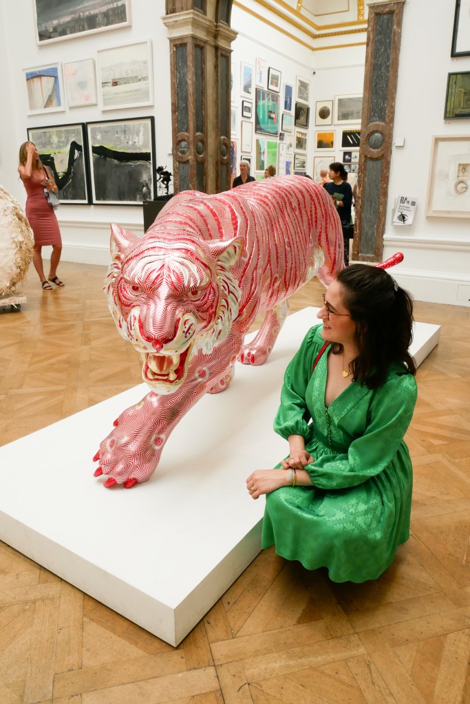 Katie is in the galleries of the Royal Academy Summer Show, where the walls and spaces are crowded with paintings, sculptures, and other artworks. She wears a green dress with long poofy sleeves, a nipped in waist and short flouncy skirt. She's carrying a red shoulder bag and a wood and blue fabric folding fan and wearing black ankle boots.
