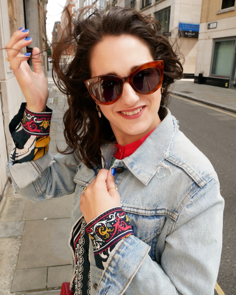 Katie wears a knee length shirt dress with multi colour print, vintage denim jacket, gold trainers and sunglasses. She smiles at the camera.