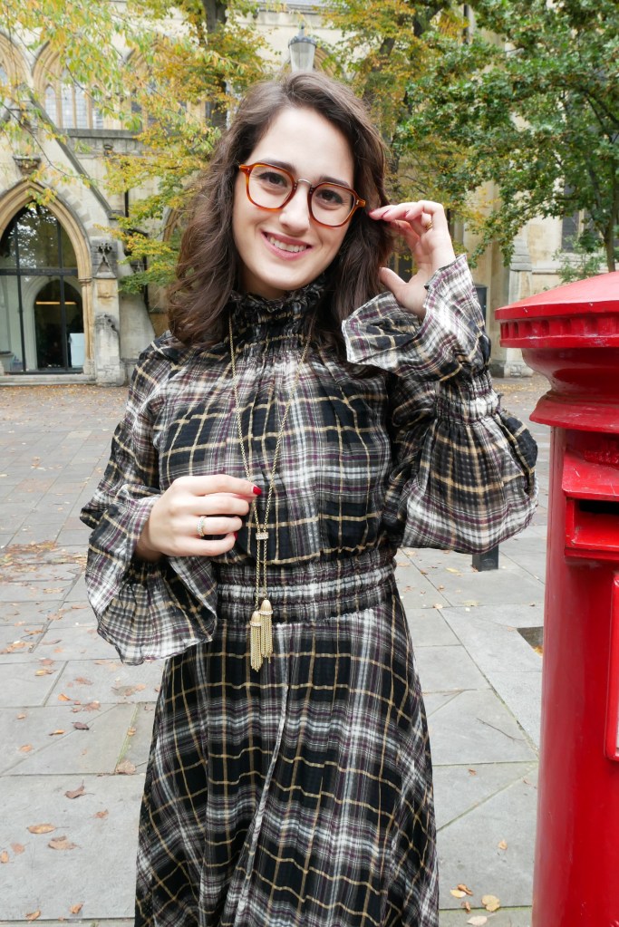 Katie wears pearl drop earrings, a plaid long sleeve dress with ruffled collar and gold tone necklace with tassels. She's walking towards the camera and her dress is swishing.