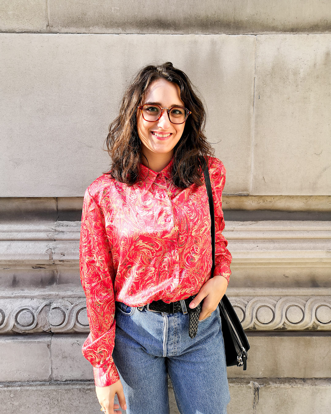 Katie wears a red and gold patterned blouse with high waisted denim cuffed above the her Dr Martens boots. Her hair is down and wavy. She's standing in front of a stone wall with minimal details.