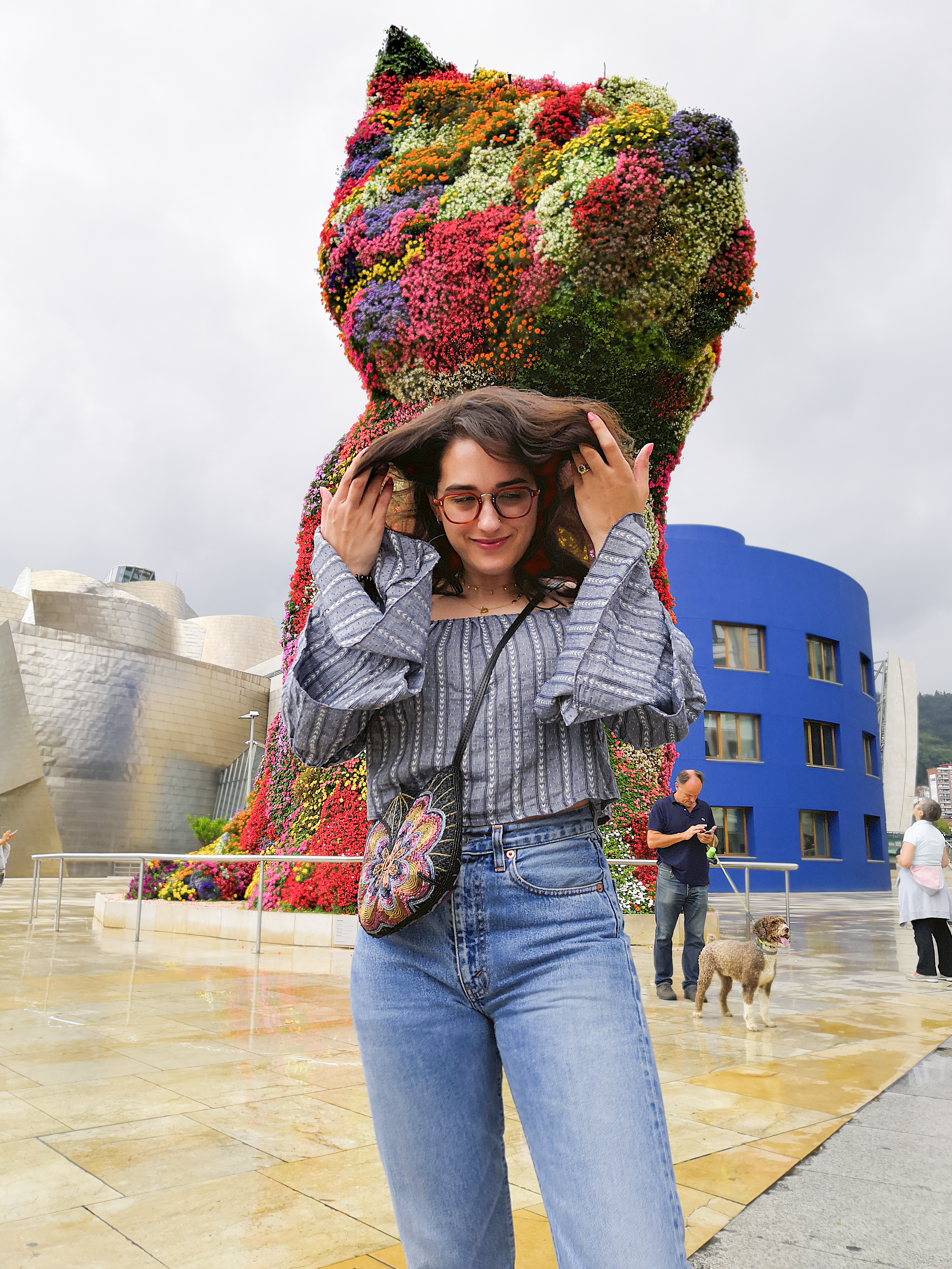 Katie is outside the Guggenheim Bilbao. She's in front of the Puppy statue. It's a drizzly day and the sky is a white / grey between showers. She wears a blue-grey off the shoulder crop top with tiered flute sleeves, high waisted jeans, white sneakers and a multicoloured beaded crossbody bag.