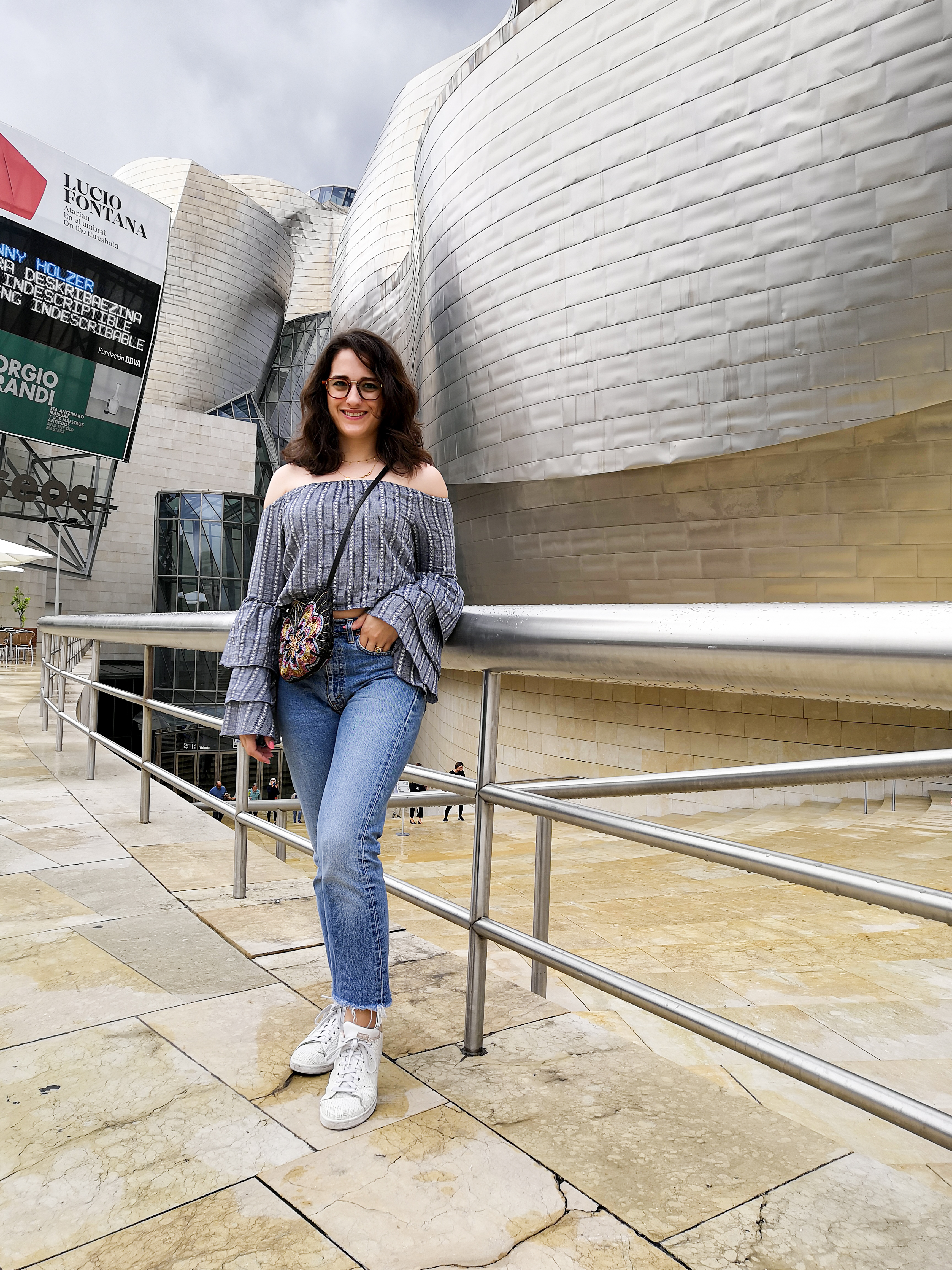 Katie is outside the Guggenheim Bilbao. She stands in front of the main entrance. It's a drizzly day and the sky is a white / grey between showers. She wears a blue-grey off the shoulder crop top with tiered flute sleeves, high waisted jeans, white sneakers and a multicoloured beaded crossbody bag.