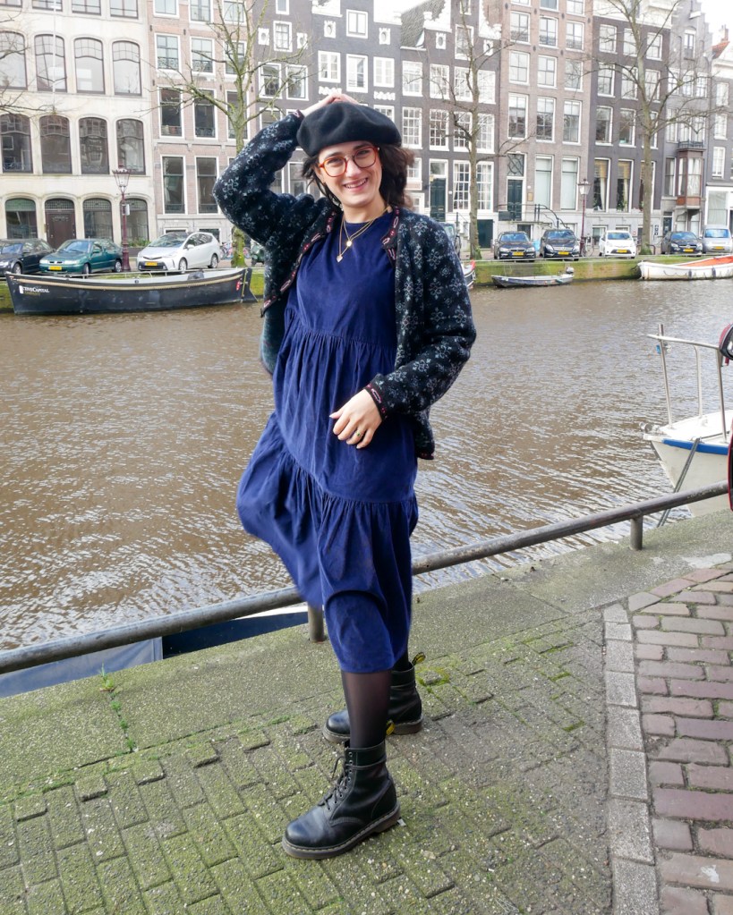 Katie stands by an Amsterdam Canal in de Pijps. She wears a black beret, billow midi navy dress, cardigan in navy and black knit, black tights and black DMs. She accessorised with a few long necklaces.