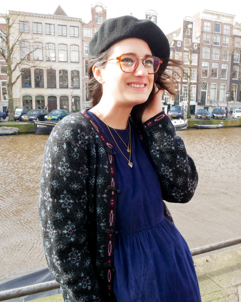 Katie stands by an Amsterdam Canal in de Pijps. She wears a black beret, billow midi navy dress, cardigan in navy and black knit, black tights and black DMs. She accessorised with a few long necklaces.