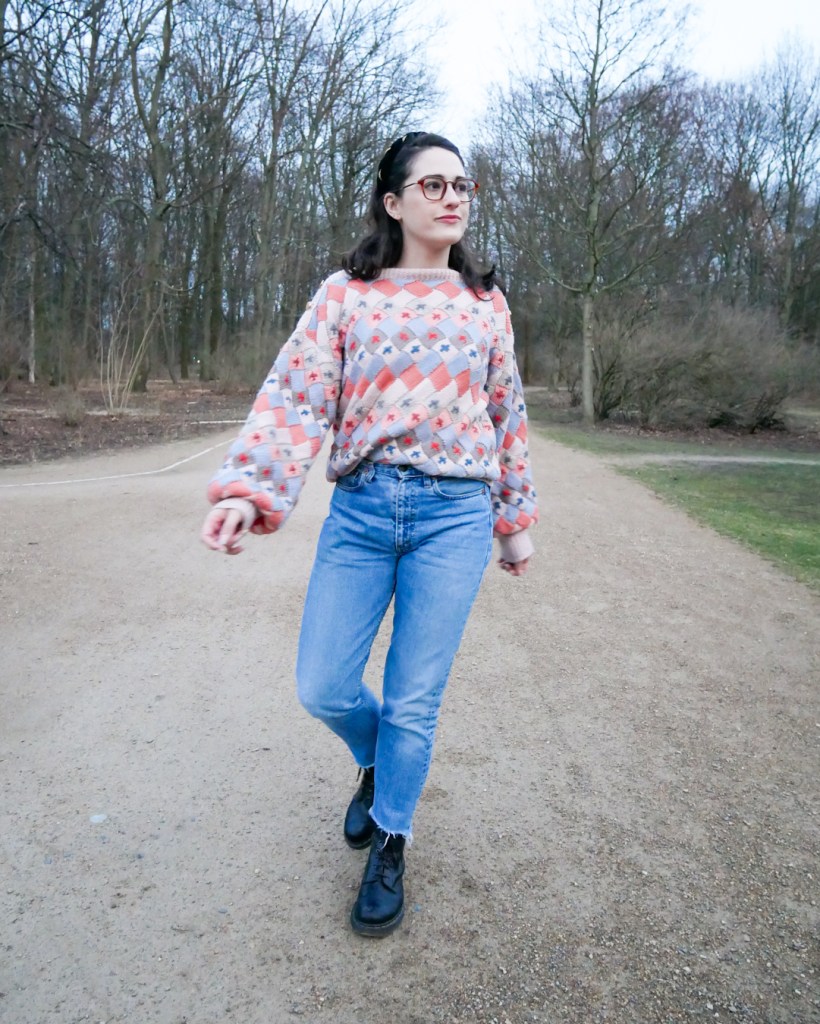 Katie wears light jeans cut off at the hems with an oversized knit 1980s style jumper in pastel colours. Her hair is pulled away from her face by a gold and black headband. She's wearing old Dr Martens boots and frolicking in a park.
