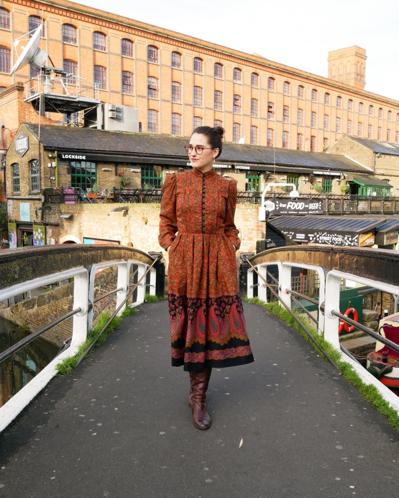 Katie wears a vintage 1970s dress with a high neck, button front to the waist, bell sleeves and A line skirt with a ditsy floral print and paisley detail at the hem in autumnal shades of orange, red, brown, blue and purple and black details.