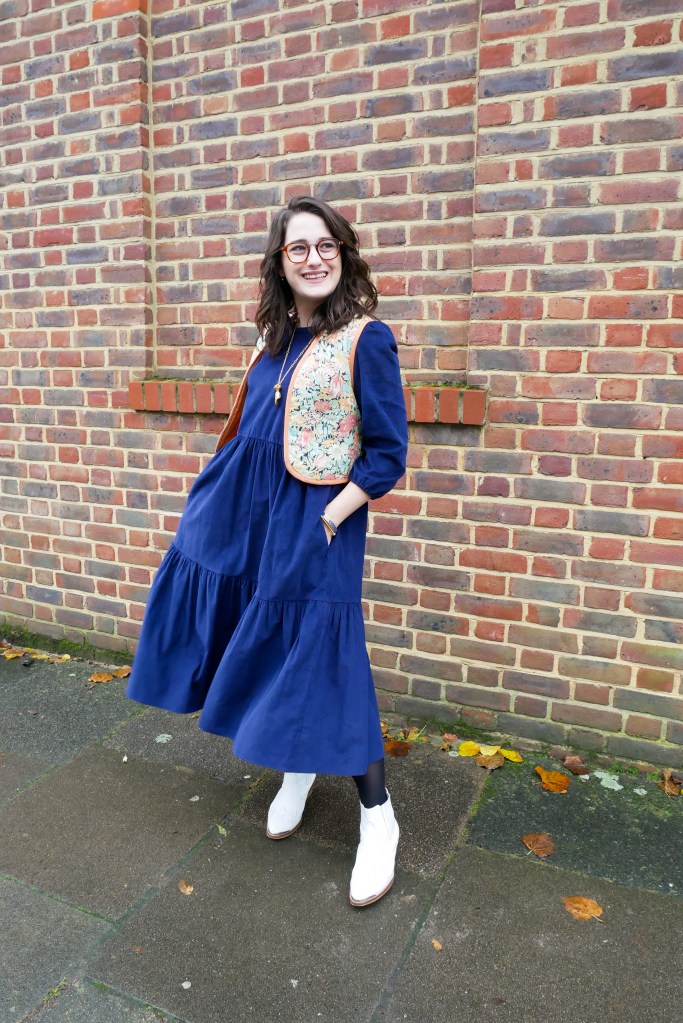 Katie wears a floaty navy courderoy midi dress with 3/4 sleeves, a vintage floral waistcoat, white boots and gold tone jewellery.