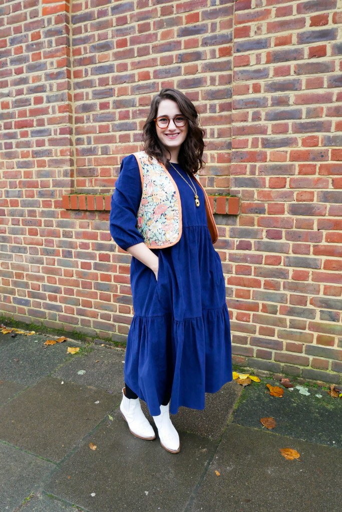 Katie wears a floaty navy courderoy midi dress with 3/4 sleeves, a vintage floral waistcoat, white boots and gold tone jewellery.