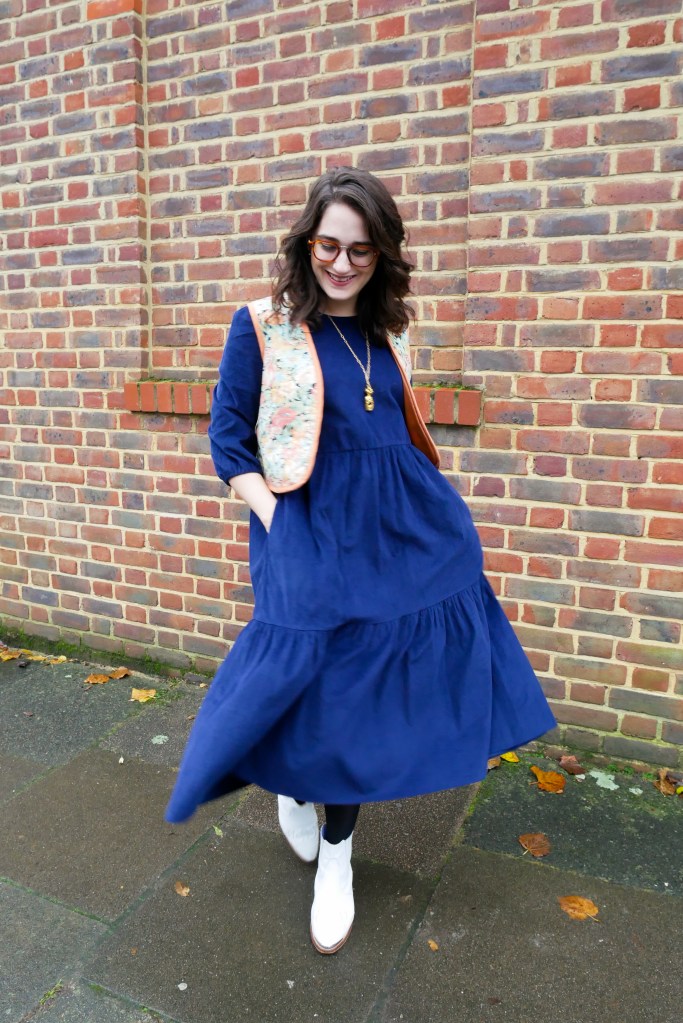 Katie wears a floaty navy courderoy midi dress with 3/4 sleeves, a vintage floral waistcoat, white boots and gold tone jewellery.