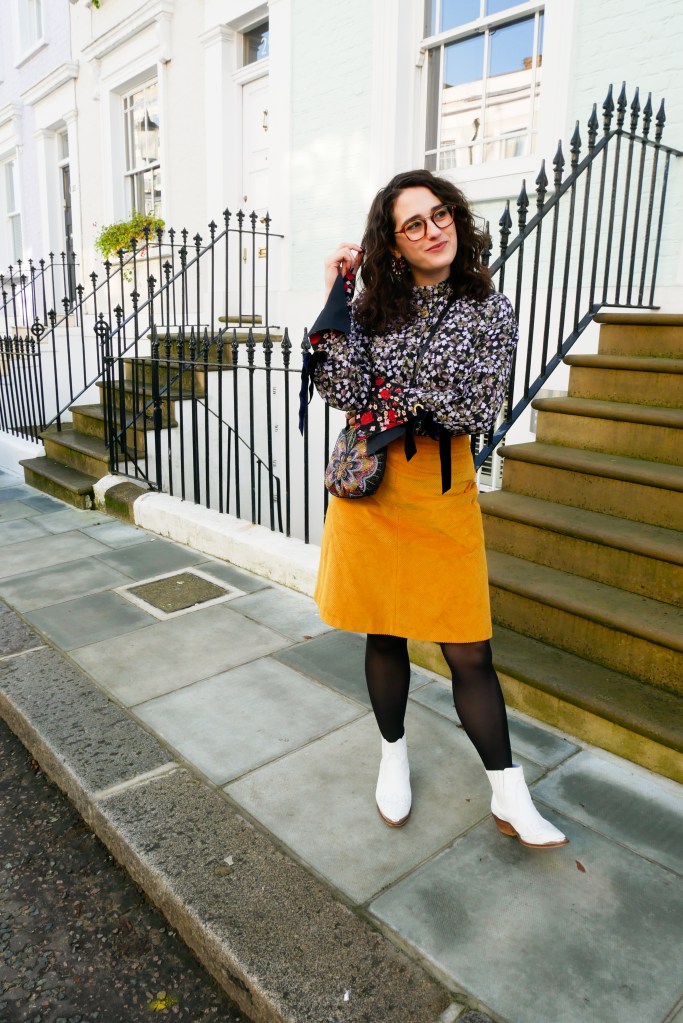 Katie wears a purple floral blouse with clashing red floral print detail at the cuffs, a mustard corduroy knee length skirt, black tights and white cowgirl style boots.