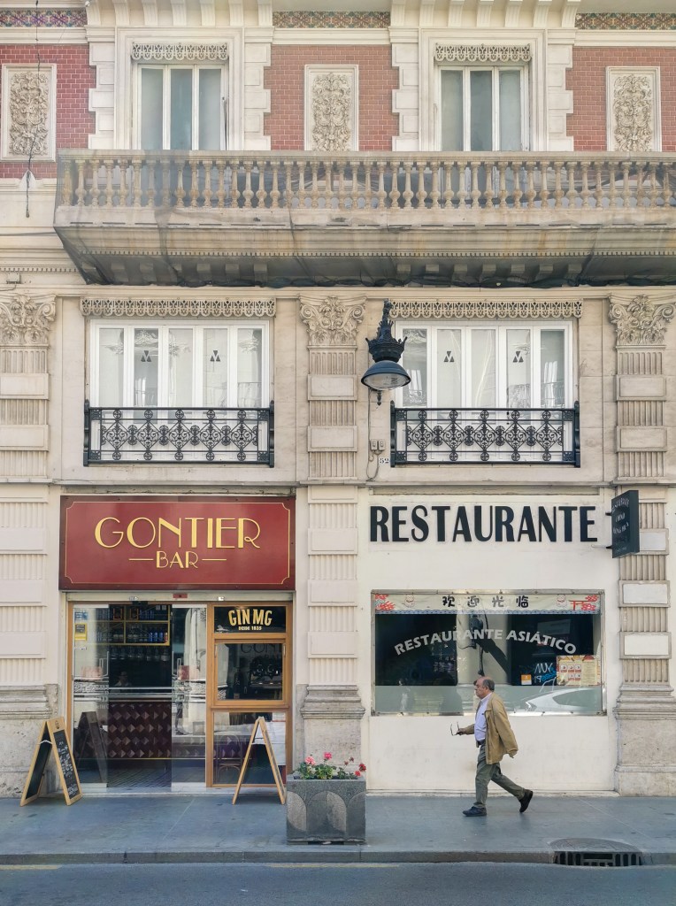 A white stone building in Valencia with two shop fronts. One shop has a large red sign that says "Gontier Bar" and the other a white sign that says "Restaurante". In front of the restaurant a man with grey trousers, a blue button down shirt, and a tan jacket walks past. In his right hand he's holding his eye glasses.