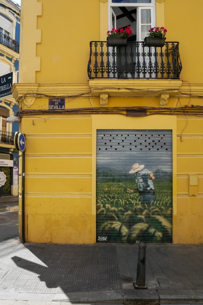 A yellow building in Valencia with a blue and white street sign saying "Calle De Sevilla". There's a shop that's shuttered, and painted on the shutters is a farmer in a field. The sky in the painting is blue with clouds, the field is a cornfield, and the farmer is wearing a white wide brimmed hat, blue button down that's rolled up to the sleeves, and blue trousers. The farmer is angled away from us so we cannot see their face.