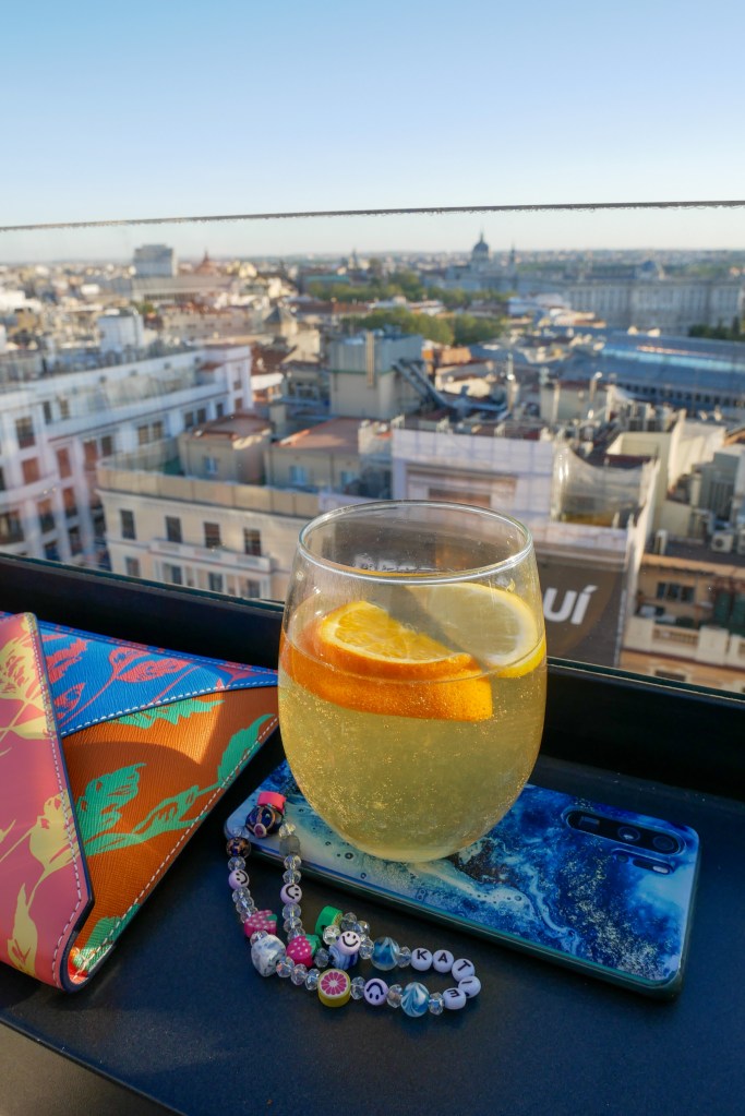 It's a bright sunny day and we are alone at a rooftop bar. We're at the ledge with a shelf for drinks, and we have an orange cocktail and mobile phone in front of us, and to the left is a multi-coloured (pink, yellow, blue, orange, green) envelope bag. We can see the skyline of Madrid in front of us.