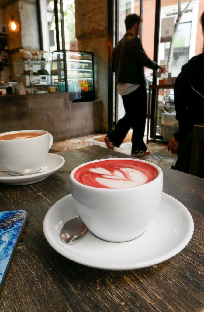 A red velvet latte in a white coffee cup on a white saucer on a wooden table. In the background is a cafe, and we can see the silhouette of someone opening the glass door to leave. Behind the person is the cafe counter with a cabinet for drinks and a display for pastries.