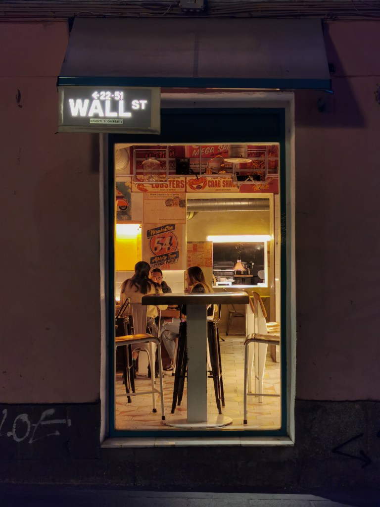 We're in Madrid at night on the street passing a bar with a small neon sign designed to look like a street sign with the text: (image of an arrow pointing left) 22-51 Wall St. The sign is above a large window where we can see into the bar. There's an empty table directly in front of us, and further back is a group of three friends engaged in conversation. The bar is bathed in a warm yellow light and except for that group, is mostly empty right now.