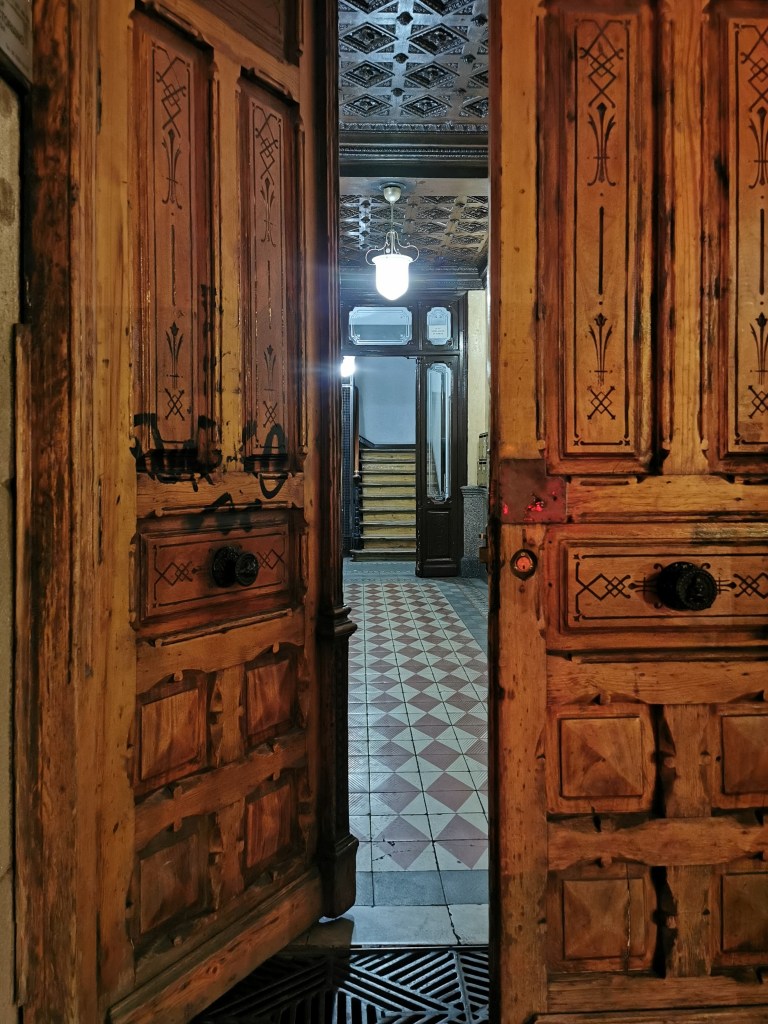 In Madrid at night we're on the street looking at a front door, made up of two heavy wooden doors with carved details and painted black geometric designs. The left door is open inwards, left open by someone else. We can see a long yellow and red tiled corridor leading to a staircase, and a large ceiling light.