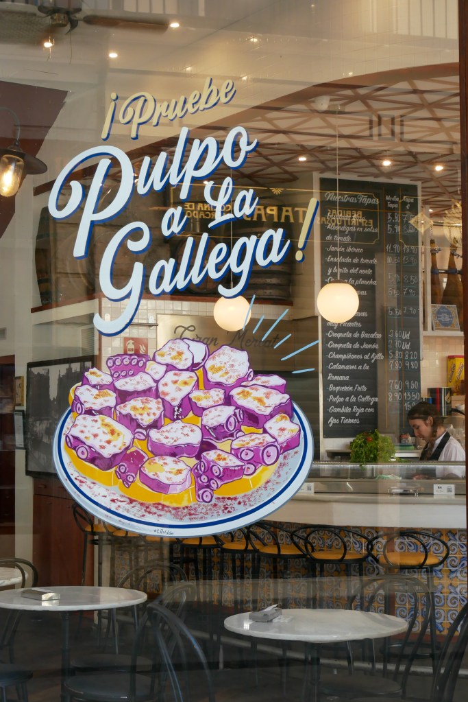 A window of a restaurant in Valencia. One the glass there is a painted picture of a plate of octopus prepared to eat. Above it in cursive are the words: "¡ Pruebe Pulpo a La Gallega!" Behind the glass we can see into the restaurant, where there's a large blackboard with menu options and a woman preparing a drink at the bar.