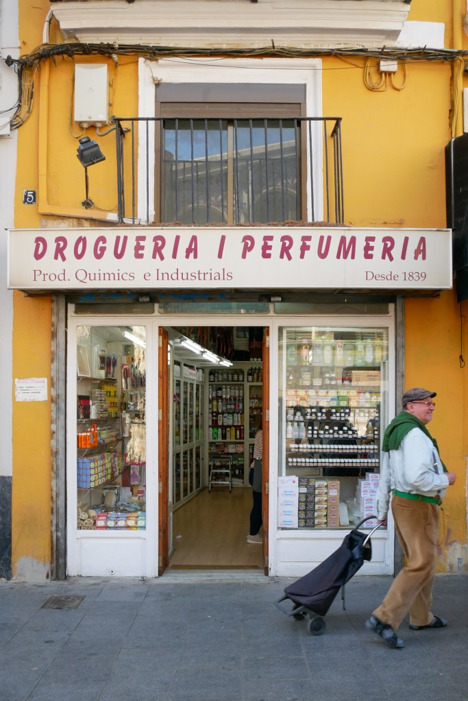 A pharmacy in Valencia. A yellow building with a white store front and white sign with red letters that read: "Drogueria / Perfumeria Prod. Quimics e Industrials Desde 1839" We can slightly see inside through an open door an glass window, shelves are filled with small bottles and boxes. A man wearing a flat cap, blue button down shirt, green sweater tied over his shoulders, and brown corduroy trousers is dragging a black shopping trolley.