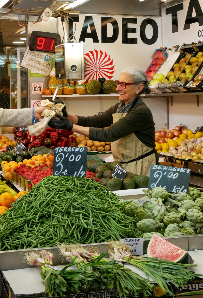 A fruit and vegetable stall in a market in in Madrid. There's a white wall in the background with a sign that says "TADEO" in black letters, and underneath it is a red circular logo. A shop worker with grey hair pulled back into a low bun wearing a dark green long sleeve top, light brown apron and thick rimmed glasses smiles as she takes a reusable bag from a customer's hand. We can only see the customer's arm, they're wearing a long sleeve grey top with thin blue trim near the wrist. Their hands meet over the strawberry display. The shop is also selling green beans, artichokes, and spring onions amongst other fruits and vegetables.