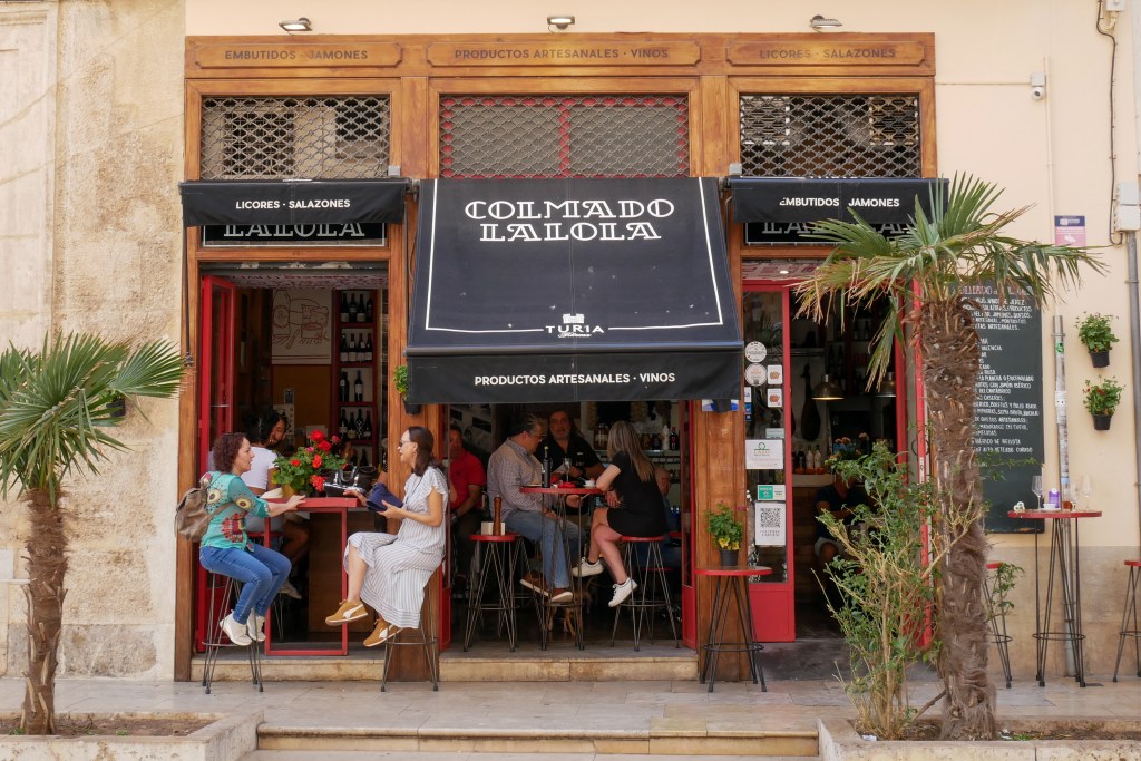 A bar in Valencia with an outdoor terrace. A large black awning with white font says "Colmado Lalola". People are sitting on high stools with high tables drinking cocktails and eating snacks.