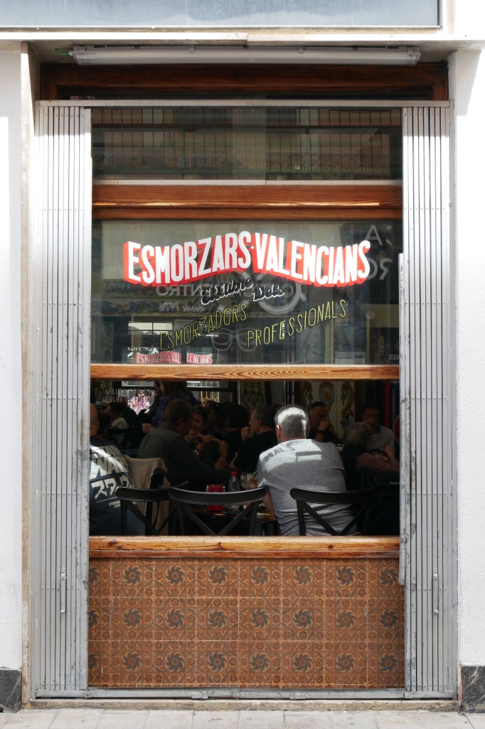 A window in Valencia. On the glass are painted red, white, and gold text saying "Esmorzars Valencians El Llibre Delo Esmorzadors Professionals" The window is half opened and we can see inside the restaurant where people are seated at tables with high-backed chairs, everyone's back is facing us as they eat and talk to each other.